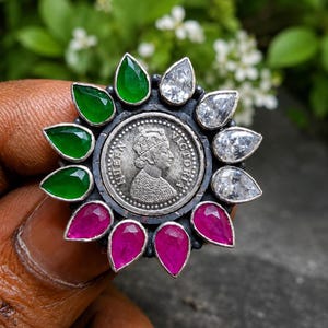 Puede incluir: Un anillo de tono plateado con una moneda central que representa a la reina Victoria con el texto QUEEN VICTORIA. La moneda está rodeada por un halo de doce piedras facetadas en forma de pera, incluyendo tres verdes, tres claras y seis rosas.