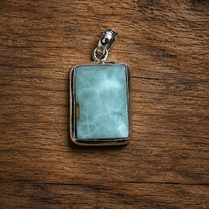 Könnte beinhalten: Rechteckiger Anhänger mit einem hellblauen Edelstein in einem silbernen Rahmen. Der Edelstein hat ein wolkiges Aussehen mit Blau- und Weißtönen. Der Anhänger hat eine silberne Öse. Schmuck.