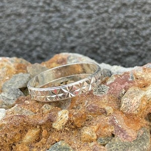 Könnte beinhalten: Ein silberner Ring mit einem sich wiederholenden Sternmuster. Der Ring wird auf einem Bett aus rauen, mehrfarbigen Steinen präsentiert. Das Design des Rings weist eine Reihe geometrischer Formen auf, die eine strukturierte Oberfläche erzeugen. Der Ring ist ein Schmuckstück.