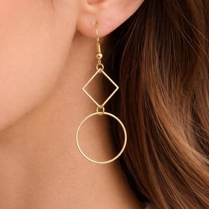 Könnte beinhalten: Goldfarbene geometrische Ohrringe. Die Ohrringe haben eine rautenförmige Form über einem kreisförmigen Reifen. Die Ohrringe hängen an einem Haken und werden vor einem Hintergrund aus braunem Haar und Haut gezeigt.