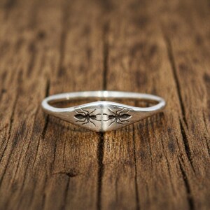 Könnte beinhalten: Ein silberner Ring mit geometrischem Design. Der Ring hat eine rautenförmige Oberfläche mit einem insektenartigen Muster. Das Band ist dünn und abgerundet. Der Ring wird auf einer Holzoberfläche gezeigt.