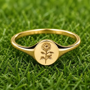 Könnte beinhalten: Ein goldfarbener Ring mit einem Sonnenblumen-Design. Der Ring hat eine kreisförmige Oberfläche mit einer eingravierten Sonnenblume und einem glatten, polierten Band. Der Ring ist vor einem Hintergrund aus grünem Gras platziert.