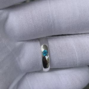 Könnte beinhalten: Ein silberner Ring mit einem einzelnen, runden, leuchtend blauen Edelstein. Der Ring hat eine glatte, polierte Oberfläche, die das Licht reflektiert. Der Edelstein ist mittig im Band gefasst und bietet ein schlichtes und elegantes Design.