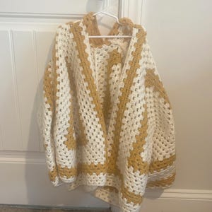 May include: A handmade, open-front crochet cardigan in a granny square pattern. The sweater features a cream-colored base with mustard yellow accents along the edges and in the square details. It hangs on a white hanger against a white door.
