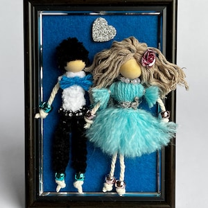 Könnte beinhalten: Gerahmtes Kunstwerk mit zwei Garnpuppen auf blauem Hintergrund. Die männliche Puppe trägt einen schwarzen Anzug mit weißem Hemd und blauer Fliege. Die weibliche Puppe trägt ein blaues Kleid. Ein Glitzerherz befindet sich oben links.