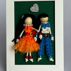 Könnte beinhalten: Ein gerahmtes Kunstwerk mit zwei Garnpuppen, die sich an den Händen halten, vor einem grünen Filzhintergrund. Die Puppe links trägt ein orangefarbenes Kleid und die Puppe rechts eine blaue Hose. Oben befindet sich ein silbernes Glitzerherz.