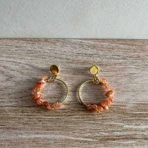Puede incluir: Un par de pendientes de aro dorados con pequeñas piedras pulidas de tonos anaranjados envueltas alrededor de la mitad inferior de cada marco circular. Cada pendiente está rematado con un perno circular dorado y liso.