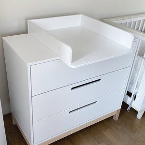 May include: A white changing table dresser with a flat top surface and a raised guard rail for changing. The unit features two wide drawers with horizontal recessed handles and is supported by light-coloured wooden legs at the base.