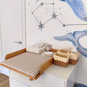 May include: A wooden changing table topper with a beige and white gingham pad, featuring a folded white blanket, a small plush sheep toy, and two woven baskets with light and dark brown textures, set against a wall with a blue whale mural.