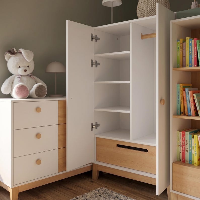 May include: A white and wood-toned nursery furniture set. The set includes a three-drawer dresser, a wardrobe with open shelves and a drawer, and a bookshelf. A stuffed bunny sits on the dresser. The wardrobe door is open, revealing shelves and a hanging rod.