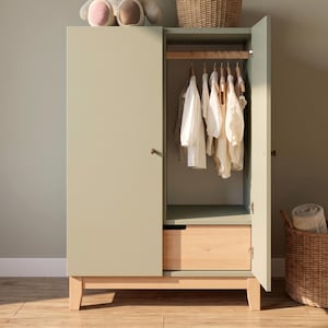 May include: A sage green wooden wardrobe with an open door revealing hanging white clothing. The wardrobe has a natural wood drawer and legs. A stuffed animal and a wicker basket are on top of and beside the wardrobe.