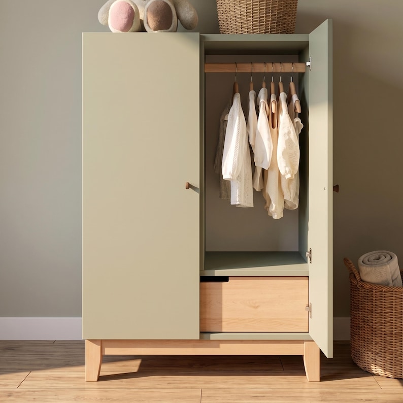 May include: A sage green wooden wardrobe with an open door revealing hanging white clothing. The wardrobe has a natural wood drawer and legs. A stuffed animal and a wicker basket are on top of and beside the wardrobe.