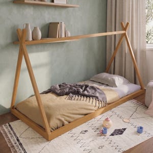 Puede incluir: Una cama de madera estilo tipi con un colch&oacute;n marr&oacute;n claro, una manta gris y una almohada blanca. La cama est&aacute; sobre una alfombra blanca con un patr&oacute;n geom&eacute;trico negro. Hay objetos decorativos en una estanter&iacute;a encima de la cama.