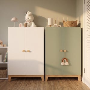 May include: Two wooden cabinets, one white and one sage green, with wooden legs and cloud-shaped handles. A stuffed bunny and a lamp sit atop the white cabinet. A woven basket and a rainbow decoration adorn the green cabinet.