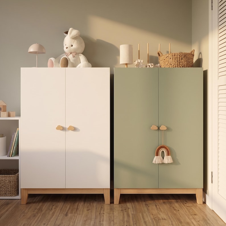 May include: Two wooden cabinets, one white and one sage green, with wooden legs and cloud-shaped handles. A stuffed bunny and a lamp sit atop the white cabinet. A woven basket and a rainbow decoration adorn the green cabinet.