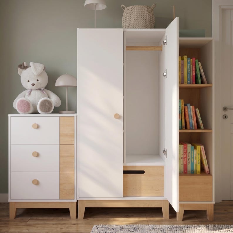 May include: A white and wood-tone nursery furniture set. The set includes a dresser with three drawers, a wardrobe with an open door, and a bookshelf filled with colorful books. A stuffed bunny sits on top of the dresser.