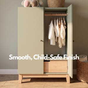 May include: A sage green wardrobe with a light wood drawer and legs. The wardrobe doors are open, revealing hanging white clothing. The text "Smooth, Child-Safe Finish" is displayed on the bottom of the wardrobe.