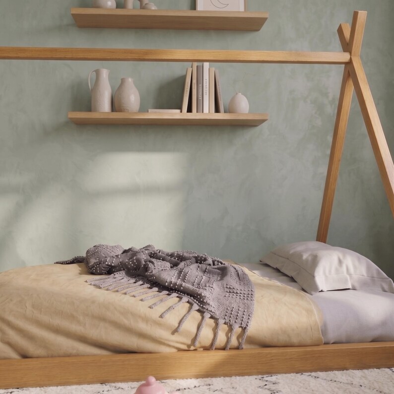 Puede incluir: Una cama con estructura de madera clara con una colcha beige y una manta gris. Una almohada blanca descansa sobre la cama. Dos estantes de madera sostienen libros y jarrones decorativos. La pared est&aacute; pintada de verde claro.