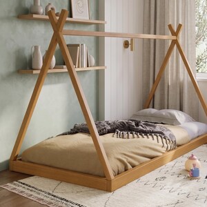 Puede incluir: Una cama de madera con forma de tipi y acabado marr&oacute;n claro. La cama tiene una colcha beige, una manta gris y una almohada blanca. La cama est&aacute; sobre una alfombra estampada en una habitaci&oacute;n con estanter&iacute;as y un aplique de pared.
