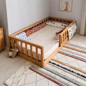 May include: A wooden toddler floor bed frame with slatted railings on three sides, featuring a light-coloured mattress, a giraffe-patterned bolster pillow, a white hanging storage pocket, and a small plush llama toy placed near the front corner.
