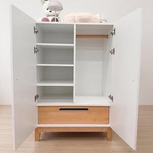 May include: White wooden wardrobe with open doors, revealing shelves, a hanging rod, and a drawer. The wardrobe has a natural wood drawer front and legs. A stuffed animal and lamp are on top.