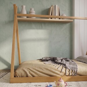 Puede incluir: Una cama con estructura de madera clara con un colch&oacute;n beige y una manta gris con flecos. La cama tiene una estanter&iacute;a encima con jarrones decorativos y libros. Un juego de t&eacute; de juguete est&aacute; en el suelo frente a la cama.