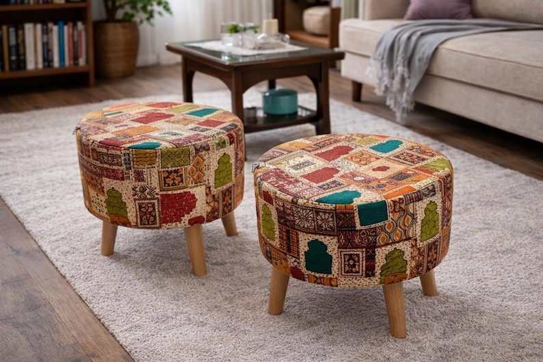 May include: Two round ottomans with a patchwork design in shades of red, orange, green, and teal. The ottomans have wooden legs and are placed on a light gray rug. The design features geometric and floral patterns.