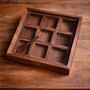 May include: A wooden spice box with nine square compartments and a small wooden spoon. The box is made of dark brown wood and sits on a wooden surface. The compartments are empty.