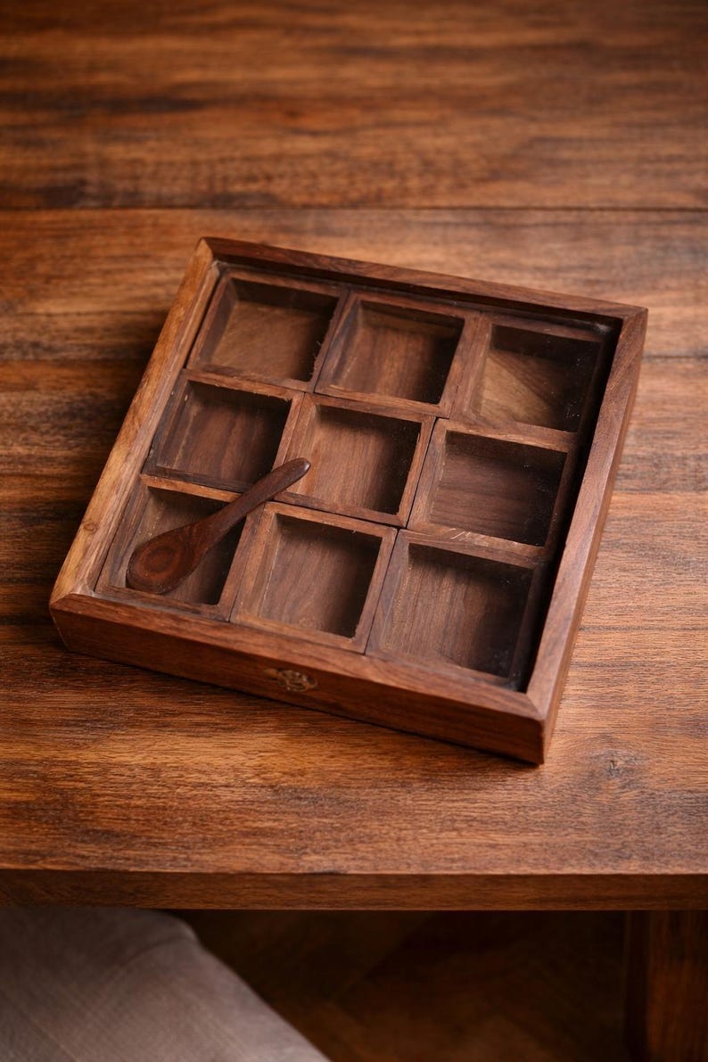 May include: A wooden spice box with nine square compartments and a small wooden spoon. The box is made of dark brown wood and sits on a wooden surface. The compartments are empty.