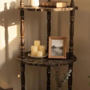 Pu&ograve; includere: Una mensola angolare in legno scuro a tre livelli con dettagli decorativi. Il ripiano superiore contiene una piccola pianta in vaso e un libro. Il ripiano centrale espone candele e una foto incorniciata. Il ripiano inferiore ha libri e un vaso di vetro.