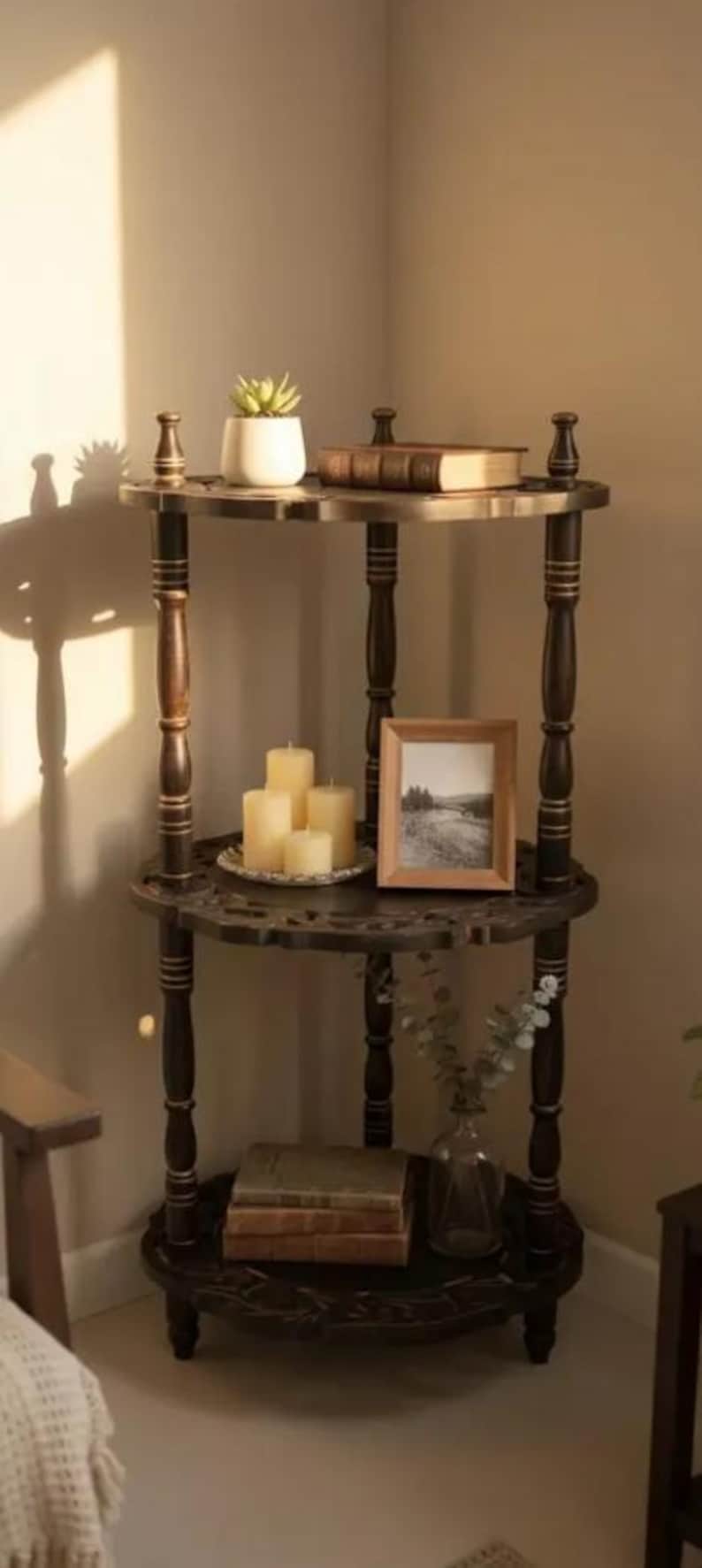Pu&ograve; includere: Una mensola angolare in legno scuro a tre livelli con dettagli decorativi. Il ripiano superiore contiene una piccola pianta in vaso e un libro. Il ripiano centrale espone candele e una foto incorniciata. Il ripiano inferiore ha libri e un vaso di vetro.