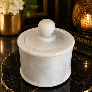 May include: A cylindrical white marble container with a matching lid featuring a spherical knob handle. The stone surface displays natural grey veining throughout the body and the lid, providing a smooth, polished finish to the vessel.
