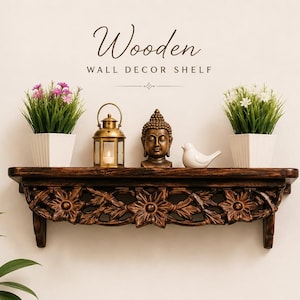 May include: A dark brown wooden wall shelf featuring intricate floral carvings along the front edge. The shelf holds two white square pots with green and pink flowers, a gold metal lantern, a Buddha head statue, and a white bird figurine.
