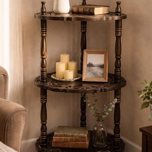 Puede incluir: Una estantería esquinera de madera oscura de tres niveles. Los estantes contienen una pequeña planta en maceta, una pila de libros, velas, una foto enmarcada y un jarrón de cristal con vegetación. La unidad tiene bordes tallados decorativos y patas torneadas.