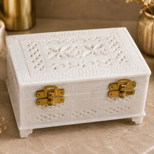 May include: A rectangular white stone box featuring intricate hand-carved lattice patterns on the lid and front panels. The box is secured with two polished gold-tone metal latches and is supported by four small, carved stone feet at the base.