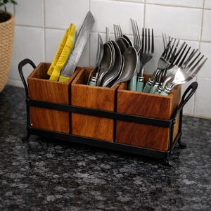 Puede incluir: Un soporte de madera para utensilios con tres compartimentos, que contiene cubiertos, pinzas y un cuchillo. El soporte tiene un marco y asas de metal negro. Los cubiertos incluyen cucharas y tenedores. El soporte est&aacute; sobre una encimera oscura.