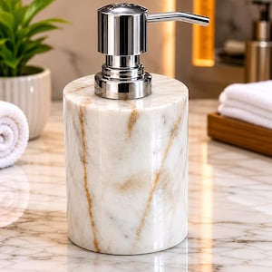 May include: A cylindrical soap dispenser made of white marble with prominent tan and gold veining. The dispenser features a polished chrome pump mechanism at the top, designed for dispensing liquid soap or lotion in a bathroom or kitchen setting.