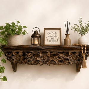 May include: A dark brown wooden wall shelf with intricate floral carvings. On the shelf sits a potted pothos plant, a bronze lantern with a lit candle, a square sign reading Collect MOMENTS not THINGS, a reed diffuser, and a beaded tassel.