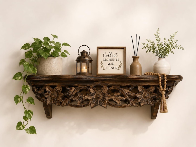 May include: A dark brown wooden wall shelf with intricate floral carvings. On the shelf sits a potted pothos plant, a bronze lantern with a lit candle, a square sign reading Collect MOMENTS not THINGS, a reed diffuser, and a beaded tassel.