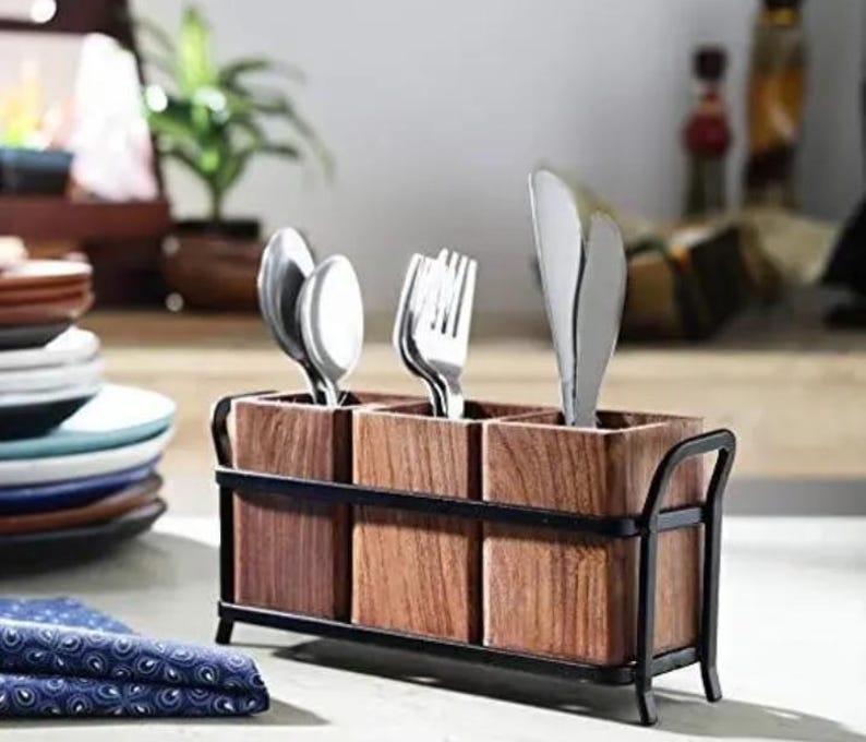 Puede incluir: Un portacubiertos de madera con tres compartimentos, cada uno con cubiertos. El soporte tiene un marco y asas de metal oscuro. La cuberter&iacute;a incluye cucharas, tenedores y un cuchillo. El soporte se encuentra sobre una superficie clara.