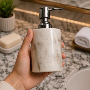 May include: A cylindrical soap dispenser made of white marble with grey veining, featuring a polished chrome pump mechanism at the top. The dispenser has a smooth, polished finish and a sturdy base, designed for dispensing liquid soap or lotion.