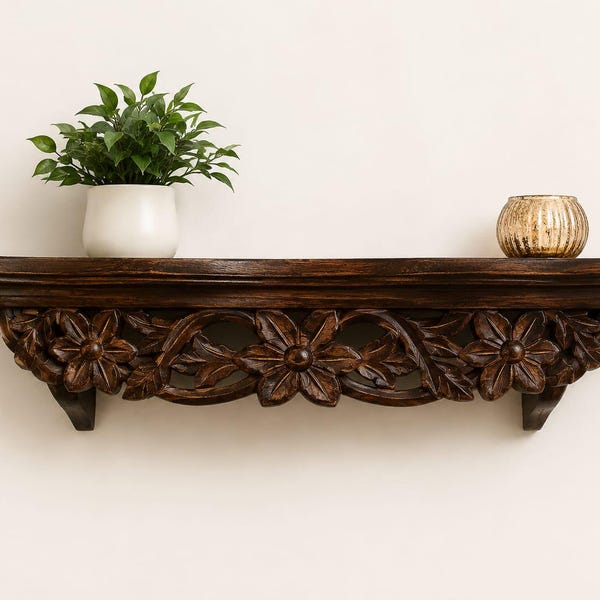 May include: A dark brown wooden wall shelf featuring intricate hand-carved floral patterns along the front edge. A white ceramic pot containing a green leafy plant sits on the left side, and a small gold-toned ribbed glass candle holder sits on the right.