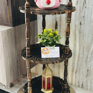 Pu&ograve; includere: Un espositore in legno marrone scuro a tre livelli con bordi intagliati. Un cestino rosa lavorato a maglia con una faccia &egrave; posto sopra. Il ripiano centrale contiene una piccola pianta e una carta con scritto "Grazie". Una lanterna decorativa &egrave; sul ripiano inferiore.
