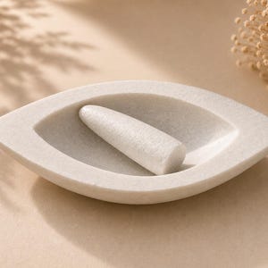May include: A white stone mortar and pestle set with an eye-shaped, almond-like bowl design. The mortar features a smooth, shallow basin, and the included pestle is cylindrical with a rounded end, both crafted from a solid, light-coloured stone material.