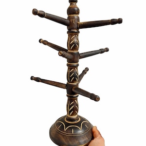 May include: A dark brown wooden jewellery display stand. The stand has multiple arms extending outwards, ideal for hanging necklaces and bracelets. The base and central column feature decorative carvings. The stand is held by a hand.