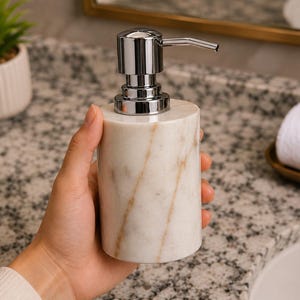 May include: A cylindrical soap dispenser made of white marble with natural tan veining. The dispenser features a polished chrome metal pump mechanism at the top. The body of the container is smooth and solid, showcasing the unique stone pattern.