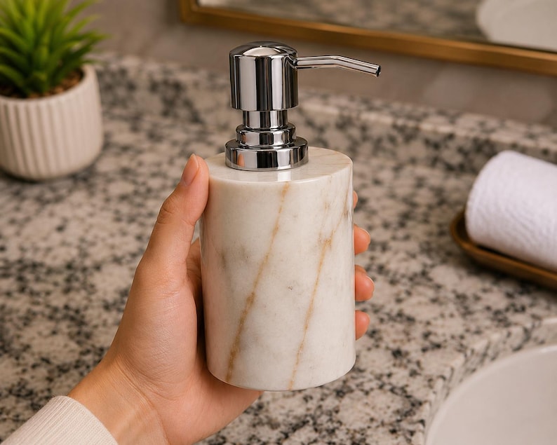 May include: A cylindrical soap dispenser made of white marble with natural tan veining. The dispenser features a polished chrome metal pump mechanism at the top. The body of the container is smooth and solid, showcasing the unique stone pattern.