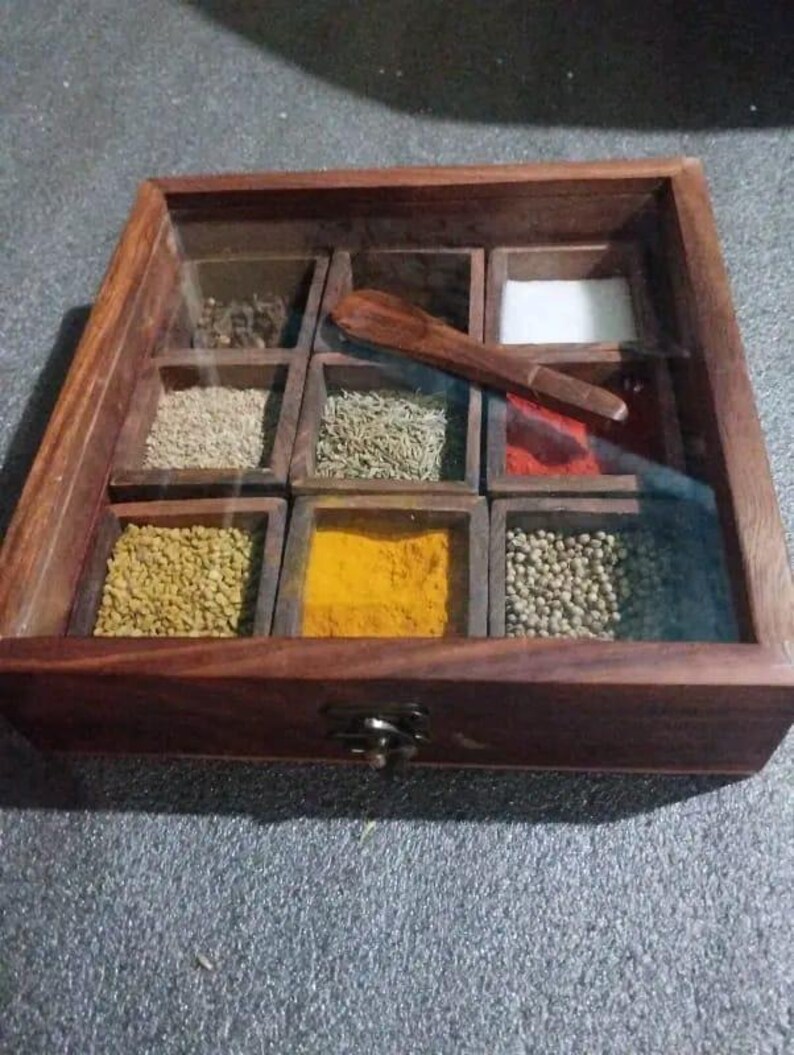 May include: A wooden spice box with a glass lid, featuring nine compartments filled with various spices and a small wooden spoon. The box is made of dark brown wood and has a metal clasp.