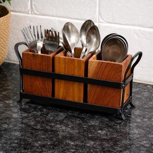 Puede incluir: Un soporte de utensilios de madera con tres compartimentos, cada uno con cubiertos. El soporte es de madera oscura y tiene un marco de metal negro con asas. Se ven tenedores, cucharas y cuchillos.