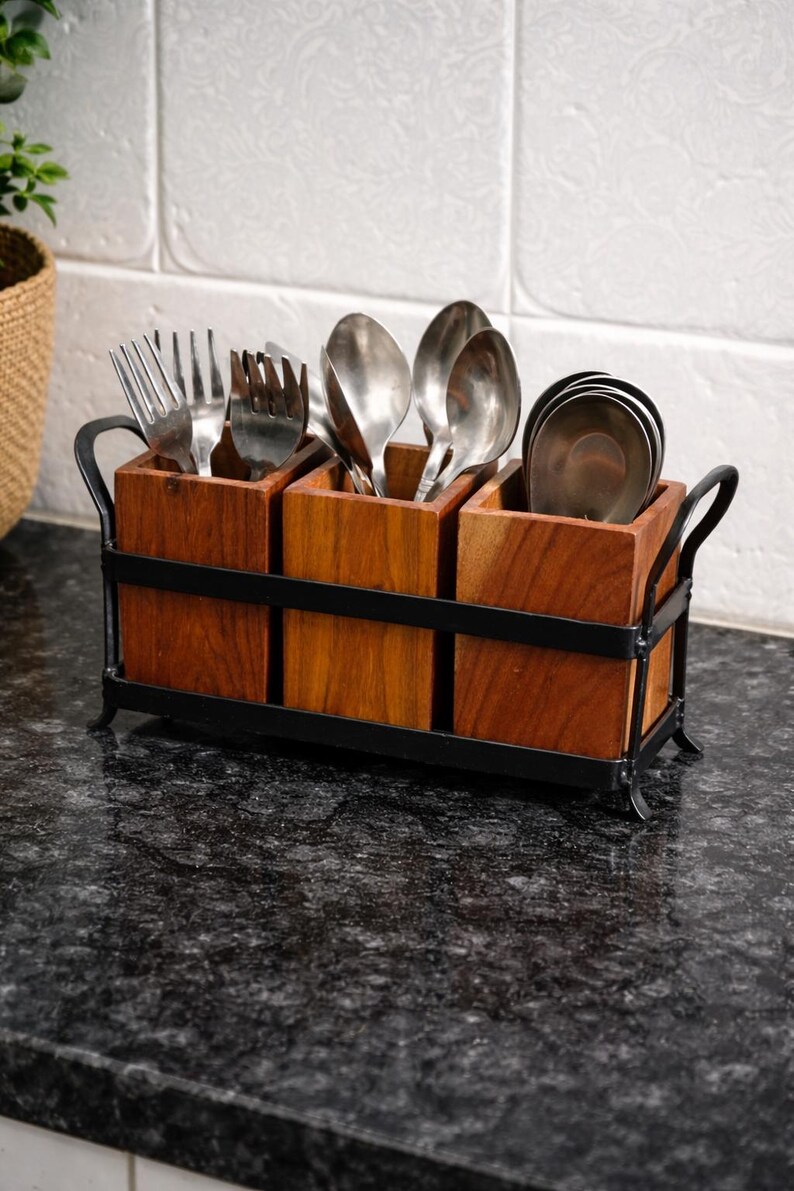 Puede incluir: Un soporte de utensilios de madera con tres compartimentos, cada uno con cubiertos. El soporte es de madera oscura y tiene un marco de metal negro con asas. Se ven tenedores, cucharas y cuchillos.